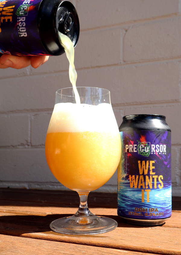 Beer being poured from a can labeled 'Pre-Cursor We Wants It' into a glass with a white brick wall background.